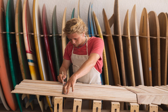 Caucasian male surfboard maker cutting wooden stripes and surfboards in the background