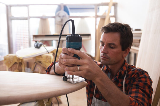Caucasian male surfboard maker shaping a wooden surfboard with a sander