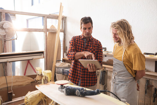 Two Caucasian male surfboard makers discussing the project and using a tablet computer - Powered by Adobe