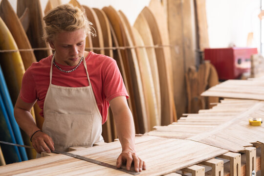 Caucasian male surfboard maker cutting wooden stripes and surfboards in the background - Powered by Adobe