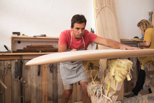 Two Caucasian male surfboard makers making a wooden surfboard together and inspecting it