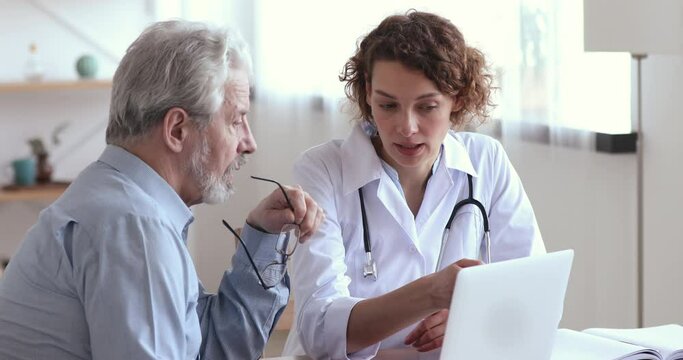 Serious young female doctor in medical uniform consulting elderly male patient, showing lab test results on computer screen at checkup visit in hospital. Focused old man listening to cardiologist.