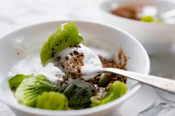 A pair of small dessert bowls with fresh kiwi fruit, white ice cream, yogurt and chocolate chips on a white stone background with teaspoons
