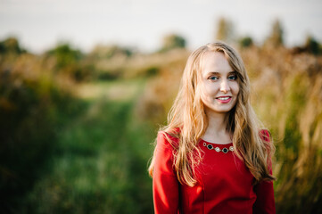 Naklejka premium Portrait of a beautiful girl stand in autumn in a red dress against the background of the field on the nature. upper half. look straight ahead. Close up.