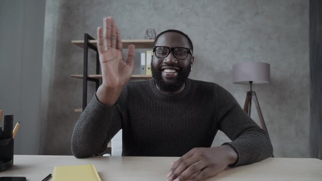 Excited African-American young guy in glasses looks into camera and greeting , waving hello. Video screen, video chat, online call, webcam shot