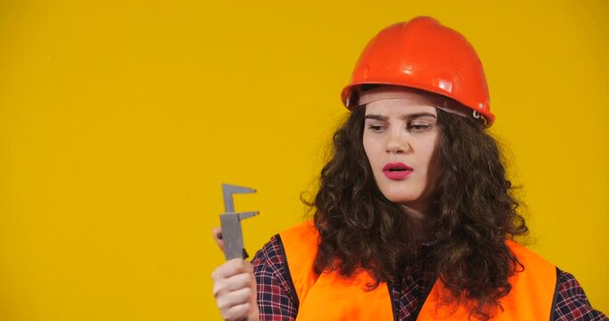 Attractive plus-size model posing. Curly-haired brunette on bright yellow studio background. Girl in red helmet and orange signal vest showing large size with caliper. Erotic Fantasy Concept