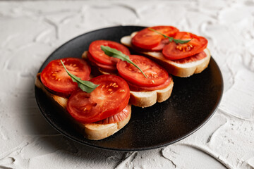 Three sandwiches with fresh and bright red tomatoes and smoked sausage, decorated with a leaf of green arugula on a matte black plate and a white stone background