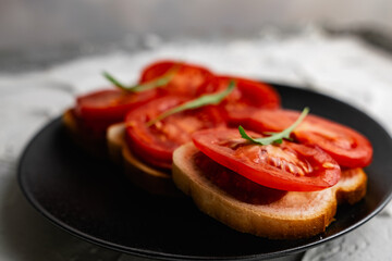 Three sandwiches with fresh and bright red tomatoes and smoked sausage, decorated with a leaf of green arugula on a matte black plate and a white stone background