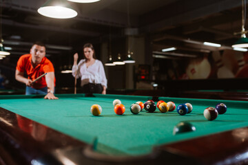 A young caucasian man in a red T-shirt is playing billiards while a girl is standing next to him. Fun time 