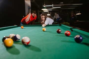 A young caucasian woman and a caucasian man play billiards. A beautiful woman is aiming a ball, while a man in a red T-shirt is standing next to her. Fun time