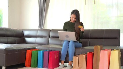 Happy asian woman shopping online using credit card and laptop computer with full colorful bags on floor in living room
