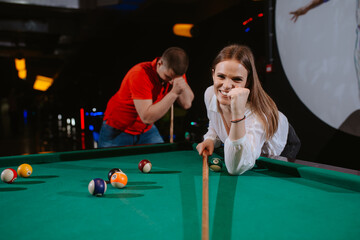 Fototapeta premium A young caucasian woman and a caucasian man play billiards. A beautiful woman is aiming a ball, while a man in a red T-shirt is standing next to her. Fun time