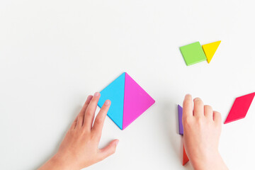 Tangram colored geometric puzzle pieces with childs hands moving pieces on white table, top view. 