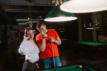 A young caucasian woman in a white shirt and a caucasian man with glasses are playing billiards and posing with sticks and balls. Fun and love