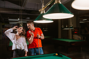 A young caucasian woman in a white shirt and a caucasian man with glasses are playing billiards and posing with sticks and balls. Fun and love