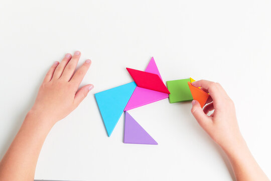 Tangram colored geometric puzzle pieces with childs hands moving pieces on white table, top view. 