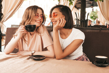 Two young beautiful smiling hipster girls in trendy summer casual clothes.Carefree women chatting in veranda terrace cafe and drinking coffee.Positive models having fun and communicating