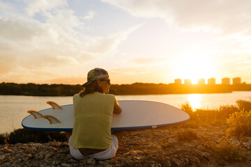 surfer sitting on a rocks by the sea one summer day at sunset He wears a cap and holds a surfboard