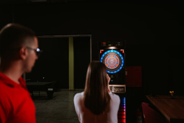 A young beautiful caucasian woman and a man are playing darts. Love and recreation