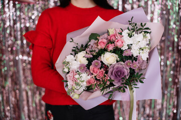 Very nice young woman holding beautiful blossoming bouquet of fresh roses, chrysanthemum, matthiola, pistachio branches, eustoma flowers in purple white and pink colors on the glittery pink background