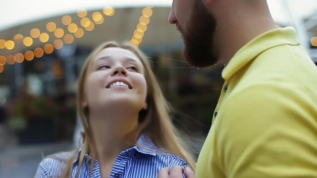 Romantic young couple in love hugs and caresses on date, blurred background of fountain and lights of restaurant outside in evening, close-up view in slow motion. Happy gy and girl resting on street.