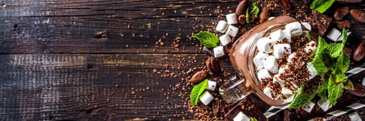 Chocolate milk shake with sauce, marshmallows and grated chocolate, decorated with mint. On old rustic wooden table with a lot of chocolate pieces, cocoa beans and chocolate shavings