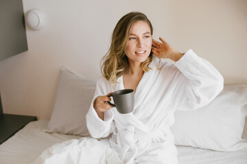 Young beautiful woman wearing white bathrobe having breakfast in bed with coffee and croissant and fresh fruits in cozy bedroom.
