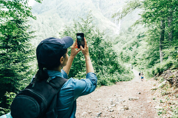 Active tourist girl with a backpack in a cap makes a specific landscape photo on the phone. Local travel in the neighborhood in nature with closed borders.