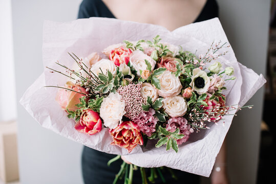 Very nice young woman holding big beautiful blossoming bouquet of fresh ranunculus, roses, carnations, tulips, ozothamnus, anemone flowers in tender white and pink colors on the grey wall background 