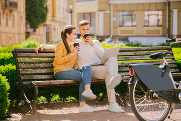 Man with laptop and woman sitting on a bench drinking coffee