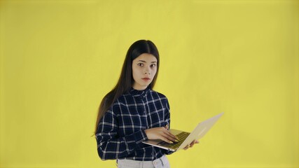 Naklejka premium Young girl with laptop looks at the camera with wide opened eyes on yellow Background in Studio