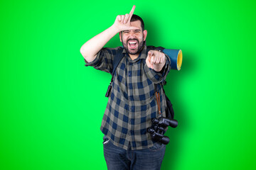 Closeup portrait serious young traveler bearded man showing loser sign on forehead looking at you with disgust at camera isolated green studio background. Negative human emotion facial expression