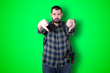 Naklejka premium Closeup portrait serious young traveler bearded man showing loser sign on forehead looking at you with disgust at camera isolated green studio background. Negative human emotion facial expression