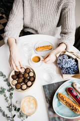 Very nice young woman sitting in a cafe, having a cup of espresso coffee coffee with some fresh eclairs, chocolate candies, eucalyptus, blue hyacinths bouquet and cappuccino cup on the white table 