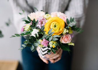 Very nice young woman holding a small beautiful blossoming flower bouquet of fresh carnations, ranunculus, matthiola in yellow and purple colors on the grey wall background