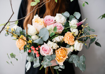 Very nice young woman holding a beautiful blossoming flower bouquet of fresh roses, eucalyptus, berries, genista in orange, white and pink colors on the grey wall background