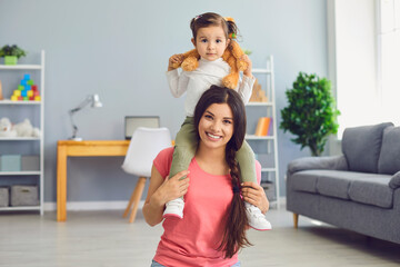 Smiling mother carrying cute little daughter on her shoulders at home. Parent and child with toy playing indoors