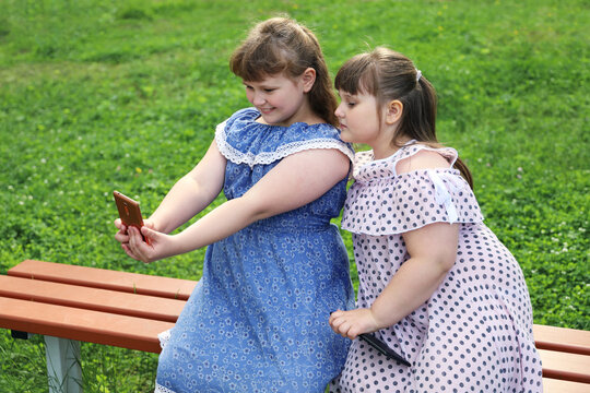 Two chubby girls take selfie on a smartphone. They are sitting on a bench on a green lawn. One is smiling and the other is sad