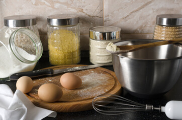 Fototapeta na wymiar Process of cooking. Closeup of wooden board, eggs, flour, glass jars with cereals, steel bowl, sugar and whisk on the black table against wall