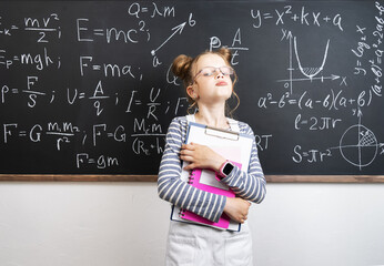 Girl schoolgirl standing at the blackboard and holding notebooks.