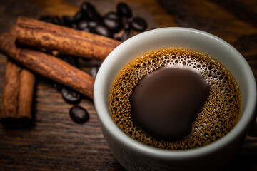 Café na xícara branca, paus de canela e grãos de café na mesa.