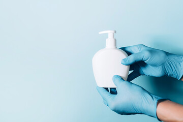 Hands in gloves holding soap bottle and medical face mask on blue background. Copy space. Preventive measures to protect against coronavirus. Products to stay safe during pandemic covid19 quarantine.