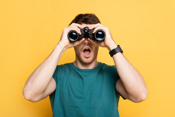 young shocked man looking through binoculars on yellow