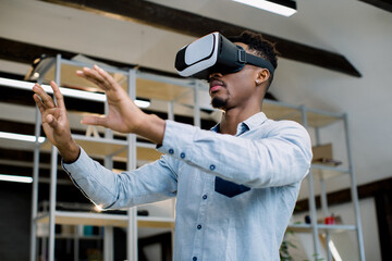 3d technology and virtual reality. Young African dark-skinned businessman dressed in casual shirt with vr goggles on head, gesturing as if interacting with something, testing video game or project