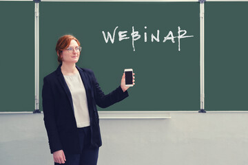 The teacher holds the phone in his hand near the blackboard with the words "Webinar", copy space. Web conferencing during isolation due to coronavirus epidemic