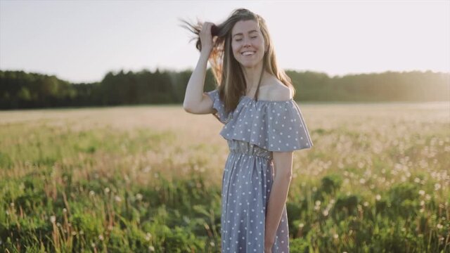 A caucasian beautiful wonan standing on the beautiful field during summer sunset and looking at the camera. The woman wears beautiful dress and. The woman is smiling. Close up. SLOW MOTION