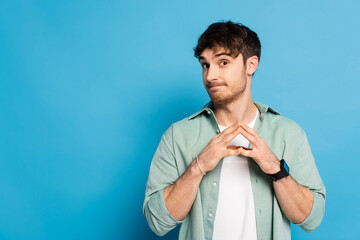sceptical young man standing with joined fingers on blue