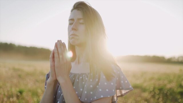 A caucasian beautiful wonan standing on the beautiful field during summer sunset and pray. The woman wears beautiful dress and. Close up. SLOW MOTION