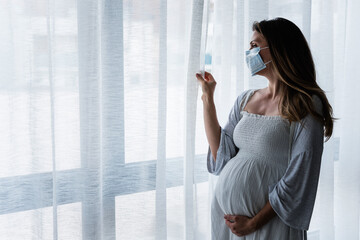 Portrait of a pregnant woman standing by the window with medical face mask, holding her belly. Worries about childbirth during pandemic.