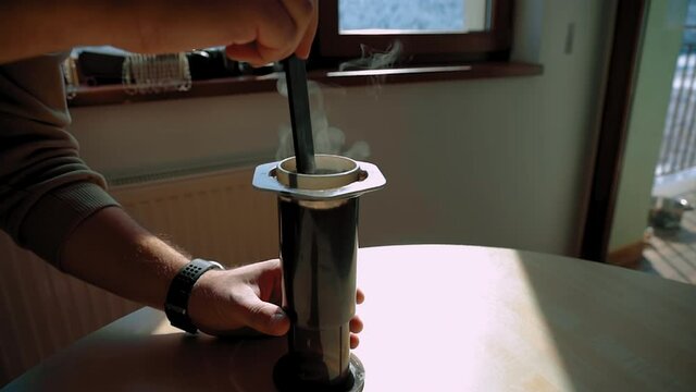 A man stirs brewing coffee in a aeropress on a wooden table